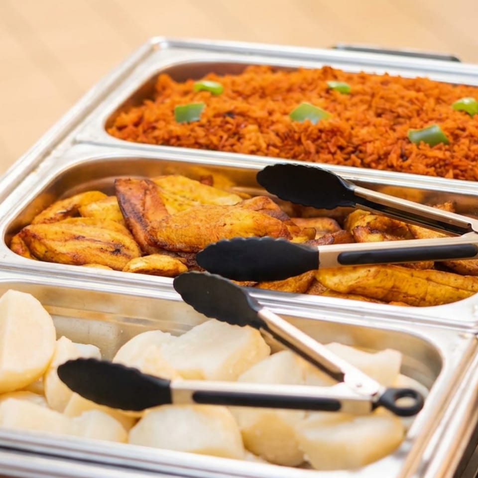 Buffet trays holding seasoned orange rice, grilled plantains, and white root vegetables with serving tongs.