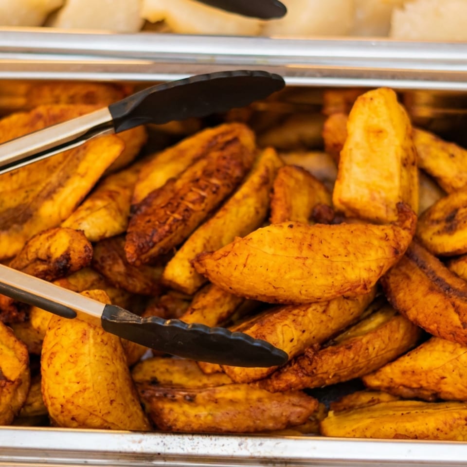 Tray of golden fried plantain slices with serving tongs in a stainless-steel buffet pan.